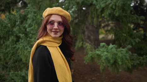 stylish woman with long red hair and painted lips, wearing a yellow beret and scarf, looking happy as she gazes at something in the background, sunglasses adding to her chic appearance in a lush park