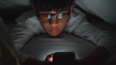 close up of young boy in bedroom at home using mobile phone to text message under covers or duvet at night