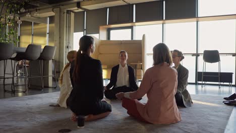 Group-of-confident-businesswomen-in-business-suits-meditate-while-sitting-on-a-carpet-in-a-modern-office.-Confident-business-girls-sitting-on-a-gray-soft-carpet-and-meditating-during-a-break-between-work-in-the-office