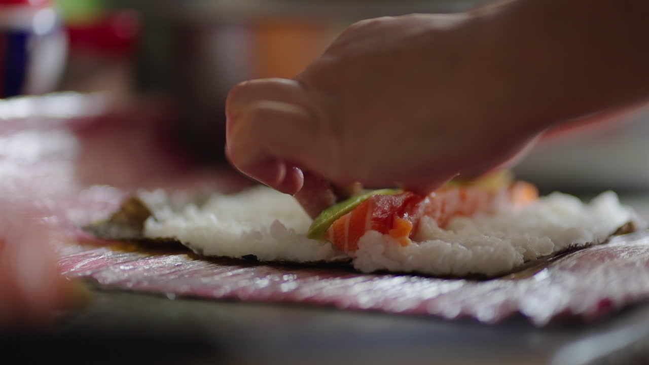 Person hand gently adds salmon - sake and avocado to vinegared rice over nori on a bamboo makisu mat, early stage of makizushi preparation in a home kitchen with blurred utensils behind, real time