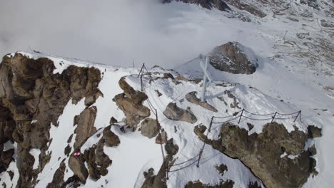 Aerial-orbiting-shot-around-summit-cross-of-snowy-Kitzsteinhorn-Mountains-in-Austria---Dense-clouds-covering-valley---top-down