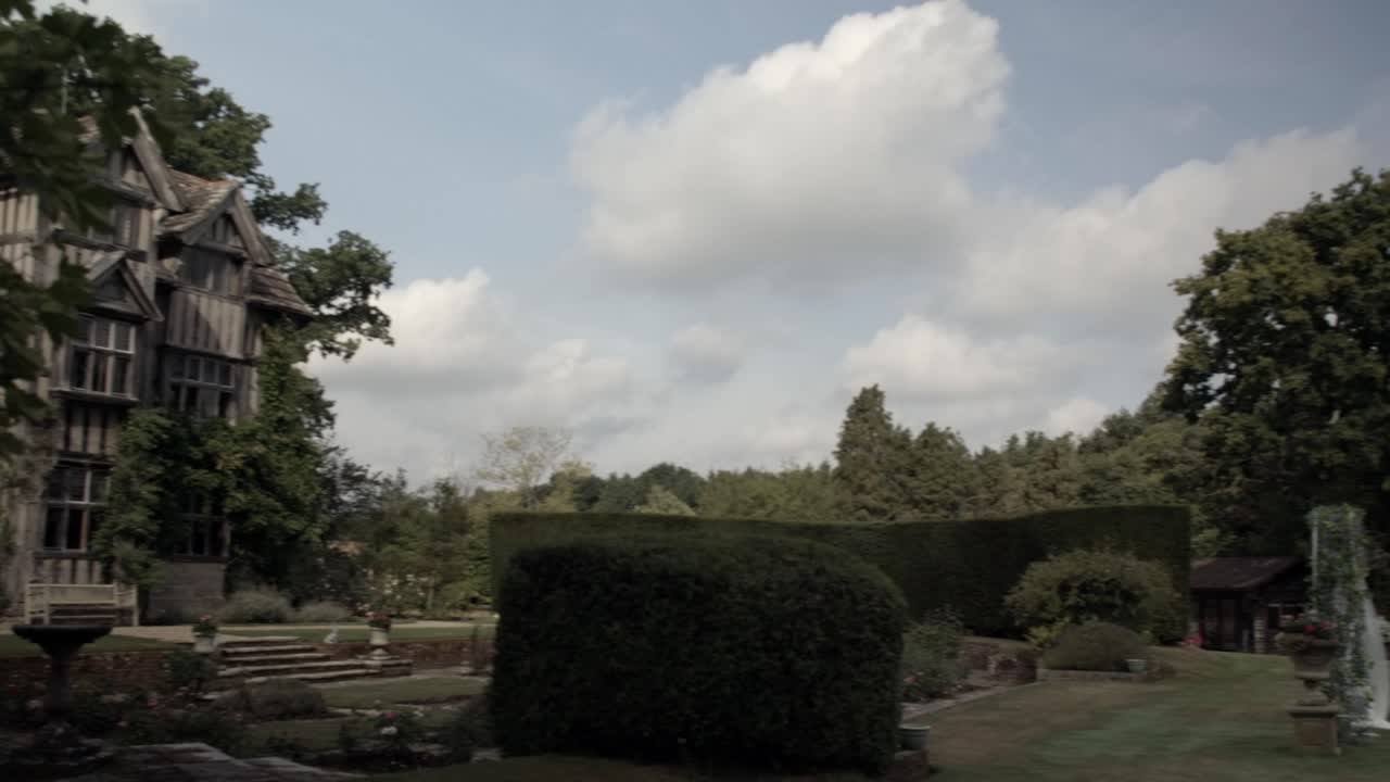 Pan shot of Tudor style heritage house peeking through garden leaves with outdoor wedding setup.
