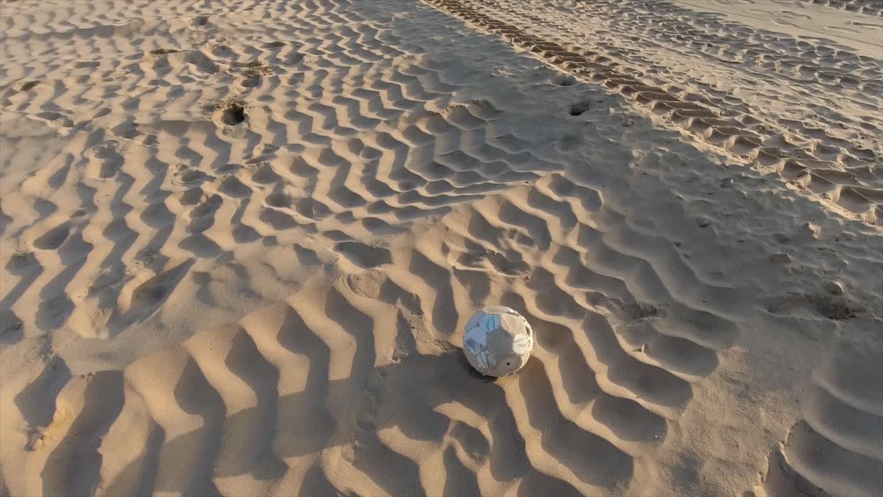 an old scruffy soccer ball rolling along beach sand with tire imprints in the sand kicked by a male with a black shoe