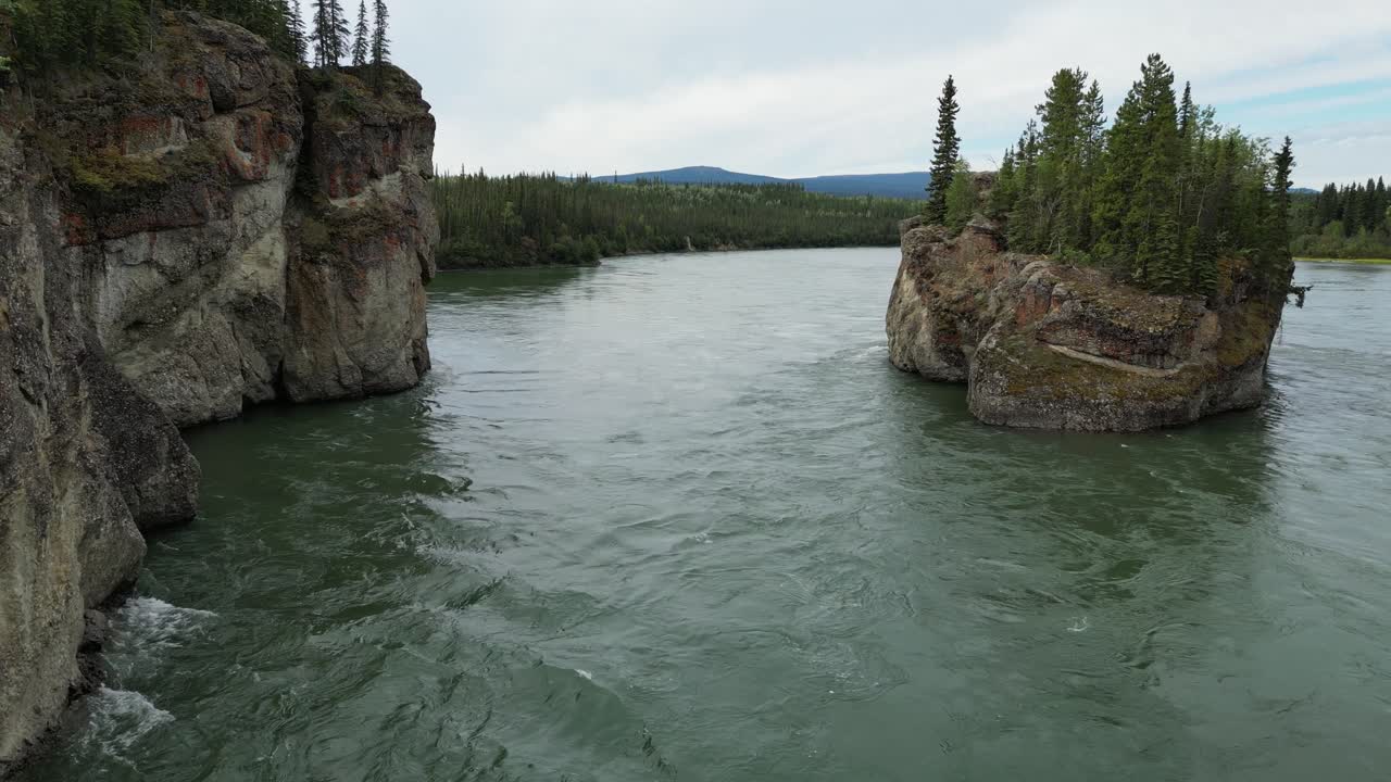 Drone dives close to the water at Five Finger Rapids in Yukon, Canada, flying between a cliffside and steep rocky island with trees, showing fast currents and visible rapids