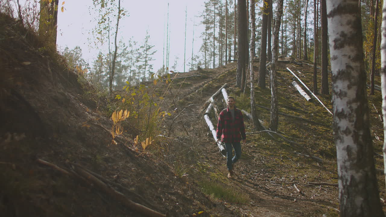 A young man walks uphill from the forest in autumn in slow motion with a backpack. A hipster man in a plaid jacket walks through a beautiful woodland area in an autumn Park. High quality 4k footage