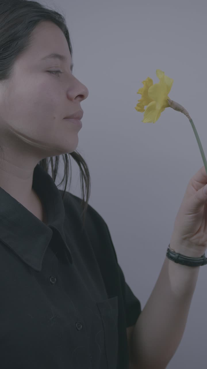 A serene vertical video of a Hispanic woman gently holding a yellow flower, eyes closed, savoring its fragrance. Soft lighting adds a calm, intimate atmosphere to the close-up shot