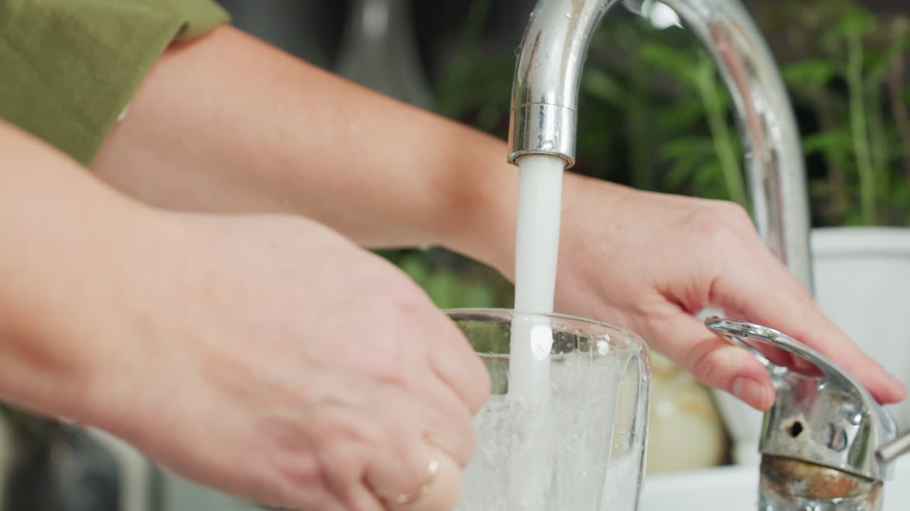 Close up of hand in green sleeve holding transparent glass under kitchen tap while filling it with water, splash visible on surface, cup about to be placed on clean counter