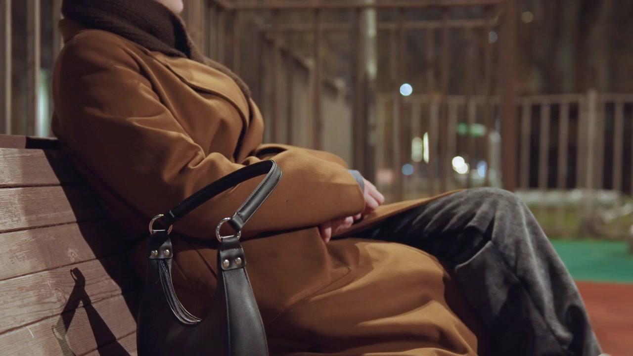 Donna caucasica che dorme su una panchina di legno di notte, cappotto marrone stretto, borsa in pelle appoggiata accanto, parco giochi vuoto e recinzione metallica sullo sfondo, bokeh morbido dei lampioni, aria autunnale fredda.