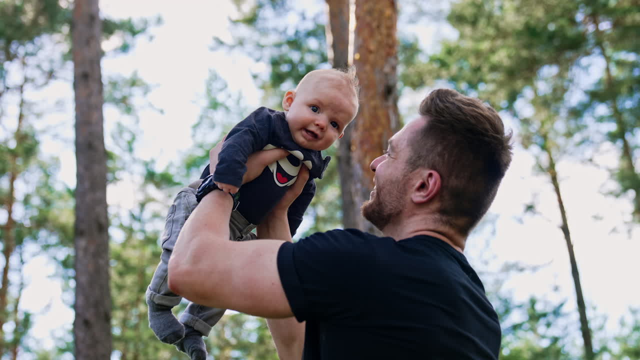 Bearded Caucasian man holding a cute newborn boy over the head. Happy dad looks with adoration on his baby. Low angle view. Pine trees at backdrop.