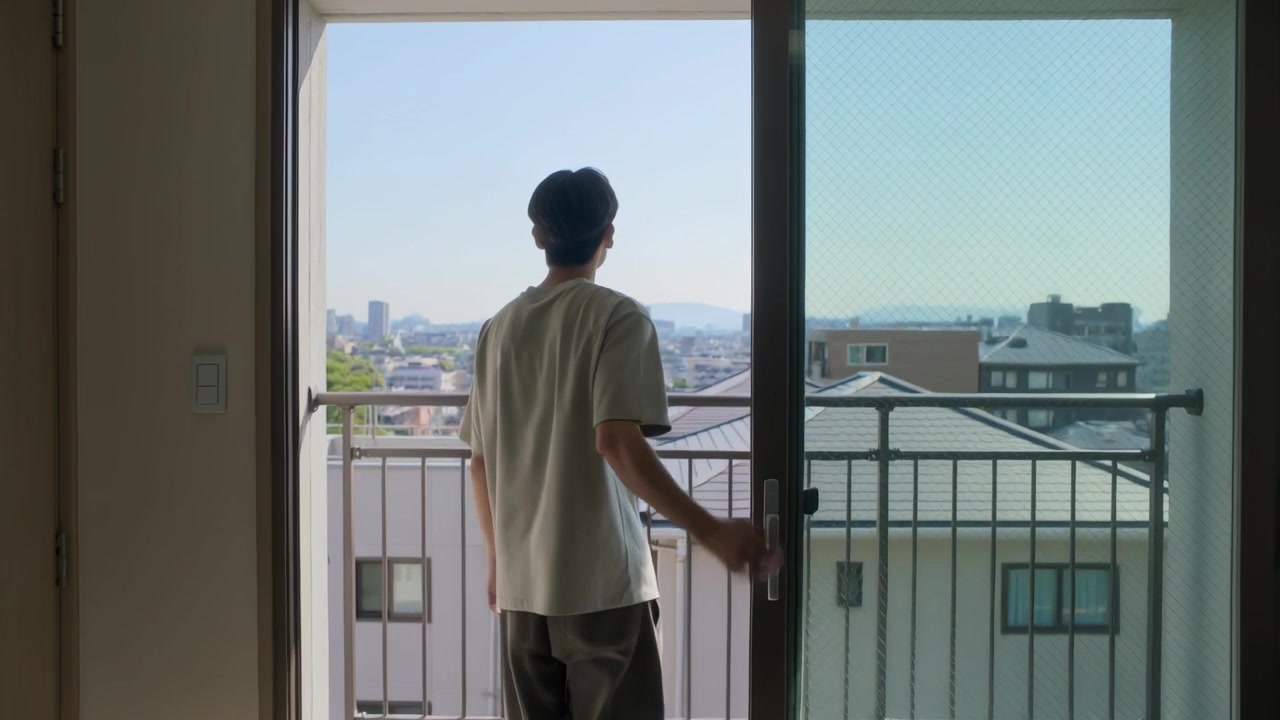 Uomo asiatico in maglietta che afferra la maniglia di una porta scorrevole, salendo sulla ringhiera di un balcone soleggiato, con vista sulla città