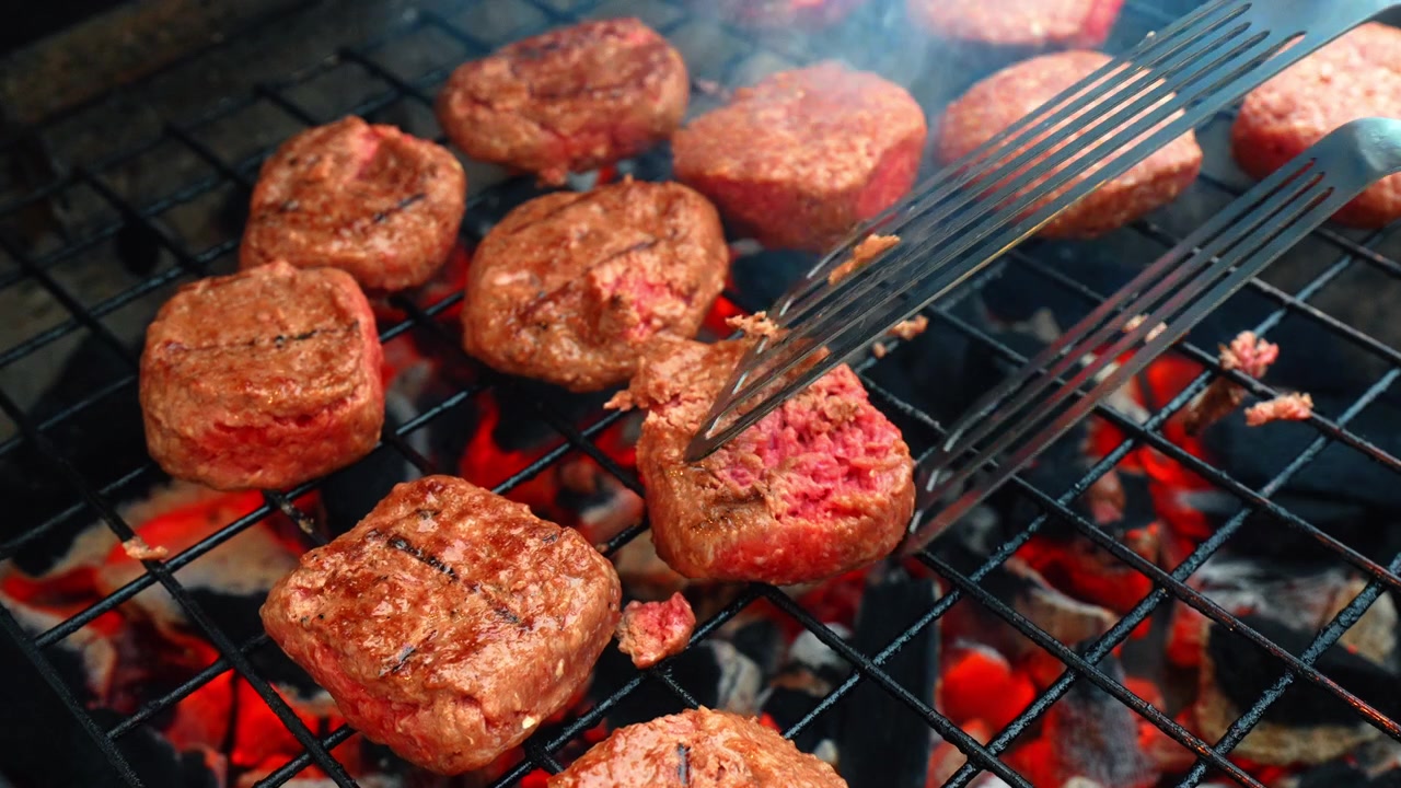 Capovolgere hamburger di carne su griglia a carbone con spatola di metallo. Scatto d'azione di una spatola di metallo che si muove sopra hamburger di carne che grigliano su carbone ardente con effetto mosso e fuoco incandescente
