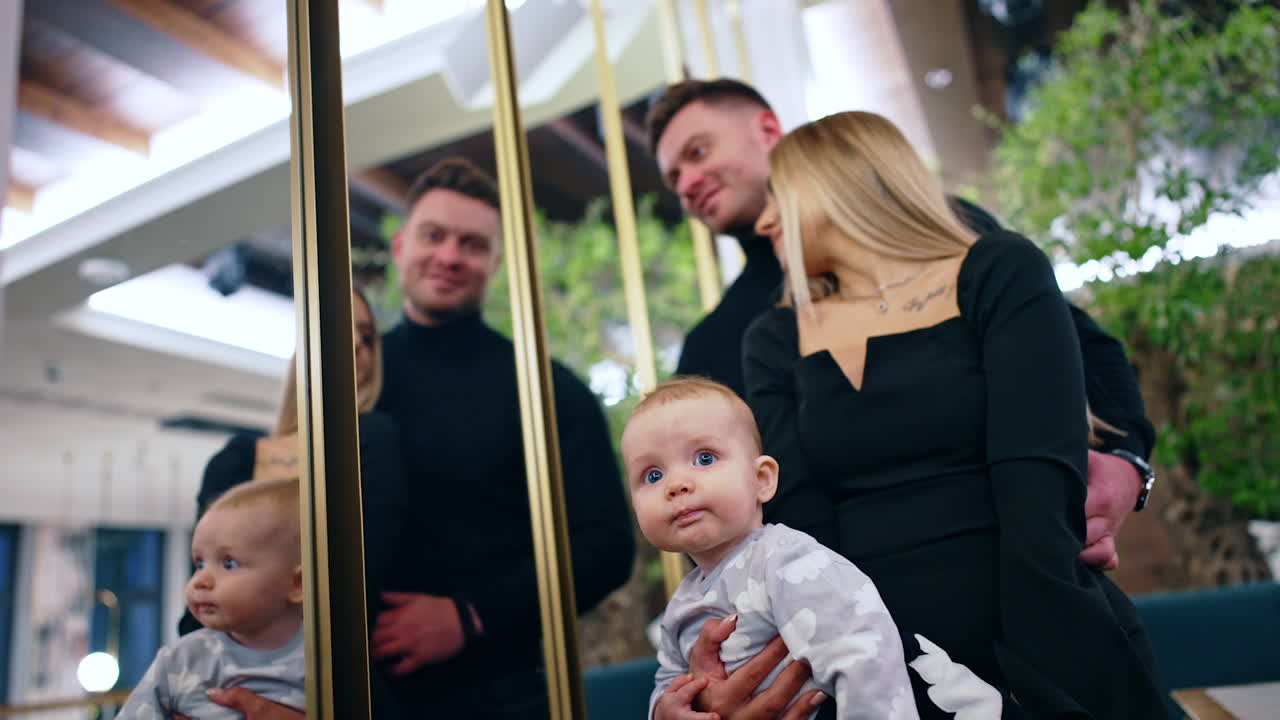 Blonde woman holds her baby boy near her belly. Smiling husband stands behind his wife looking into the mirror. Low angle view.