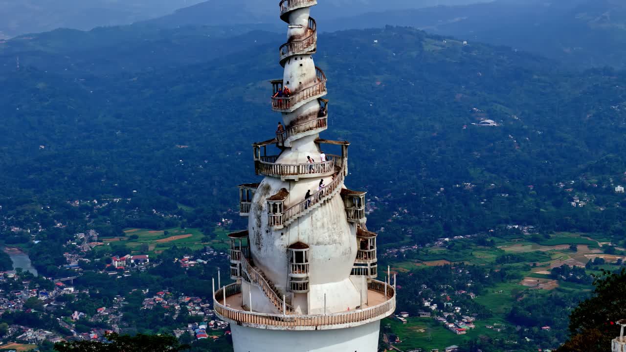 Nærbillede af hvide spiral Ambuluwawa Tower, der rejser sig over frodige bakker og fjerne bjerge i Sri Lanka, optaget af drone med en blå himmelbaggrund