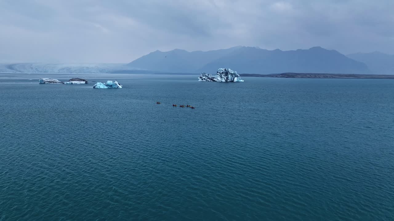 Sakin bir buzul gölünde, mavi buzdağları ve hafif yağmur bulutları altındaki katmanlı dağ sıralarının önünden geçen küçük bir grup kayakçının drone ile havadan çekimi