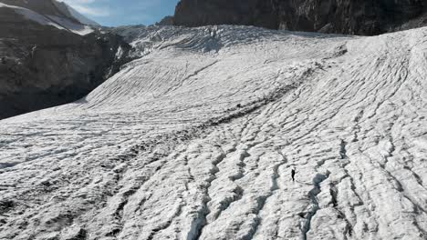 Aerial-flyover-over-a-hiker-walking-across-the-ice-of-the-Allalin-glacier-near-Saas-Fee-in-Valais,-Switzerland-as-the-sun-shines-from-behind-the-peaks-of-the-Swiss-Alps