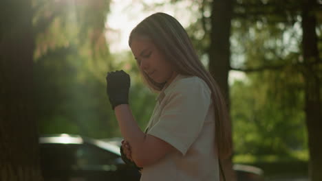 young adult wearing black gloves rubs elbow with sunlight filtering through leaves, casting a warm glow on her, blurred background with moving car