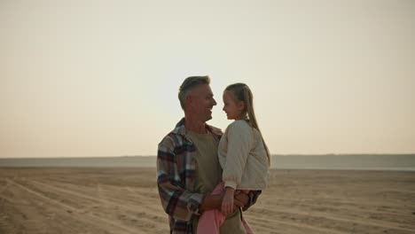 A-happy-middle-aged-brunette-man-with-gray-hair-in-a-checkered-shirt-walks-along-a-deserted-seashore-and-carries-in-his-arms-his-little-daughter,-a-blonde-girl-in-a-white-jacket,-during-his-hike-and-relaxation-outside-the-city-in-the-summer-evening