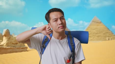close up of asian male hiker with mountaineering backpack showing thumbs down gesture while traveling in pyramid of giza