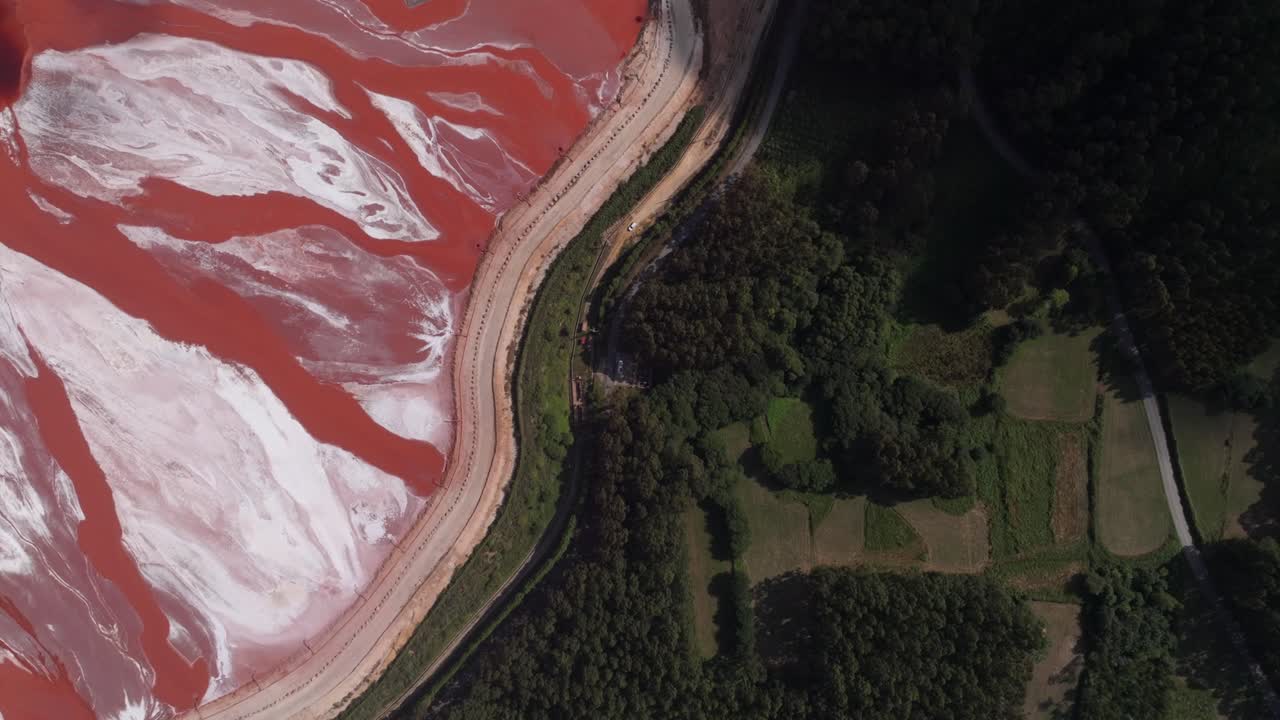 Vista aérea de uma enorme lagoa de lama vermelha na costa, um subproduto de resíduos industriais da produção de alumina. A imagem destaca a enorme escala do reservatório, com fortes contrastes entre o r
