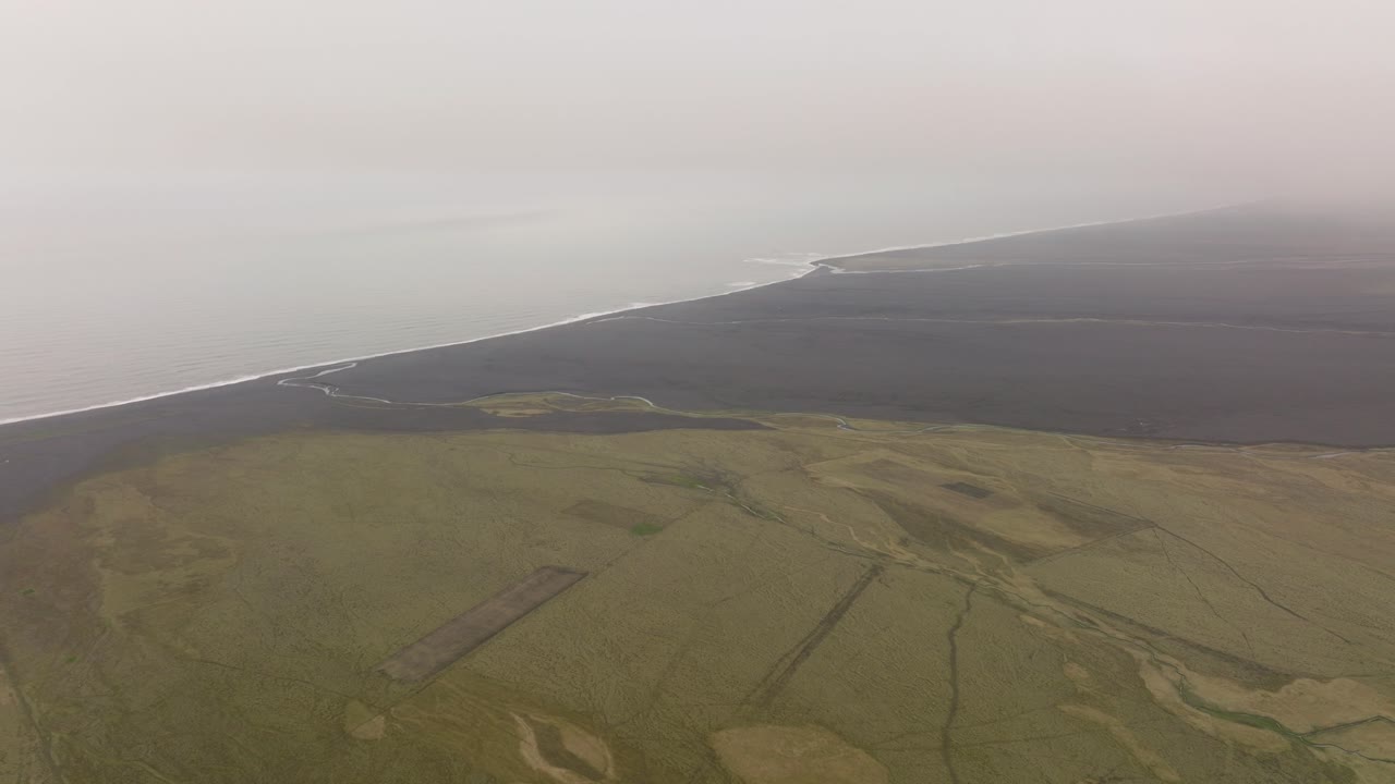 Aerial shot of coastal plains near Petursey, Iceland, under a misty sky, featuring a blend of farmland and black sand beaches stretching toward the ocean.