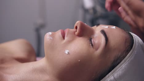 closeup view of a woman with her eyes closed lying on the couch while professional cosmetologist is applying face cream and making face massage in spa salon