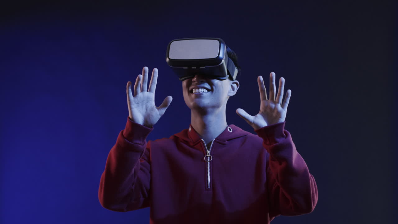 Asian man using vr headset, touching virtual screen on blue background, slow motion
