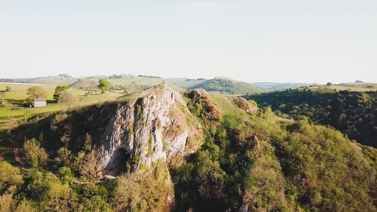Aerial drone shot moving away from Thor's Cave, Ashbourne, Peak District at sunset. Wide shot showing the cave and green rolling fields, forests, valley and clear skies.
