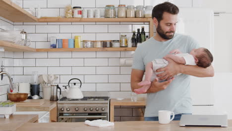 Multitasking-Vater-Hält-Seinen-Schlafenden-Kleinen-Sohn,-Während-Er-In-Der-Küche-Putzt-Und-Am-Laptop-Arbeitet