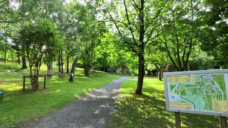a steady camera walk along a gravel path through lush, sunlit botanical gardens, ending at a detailed informational map surrounded by green trees and benches