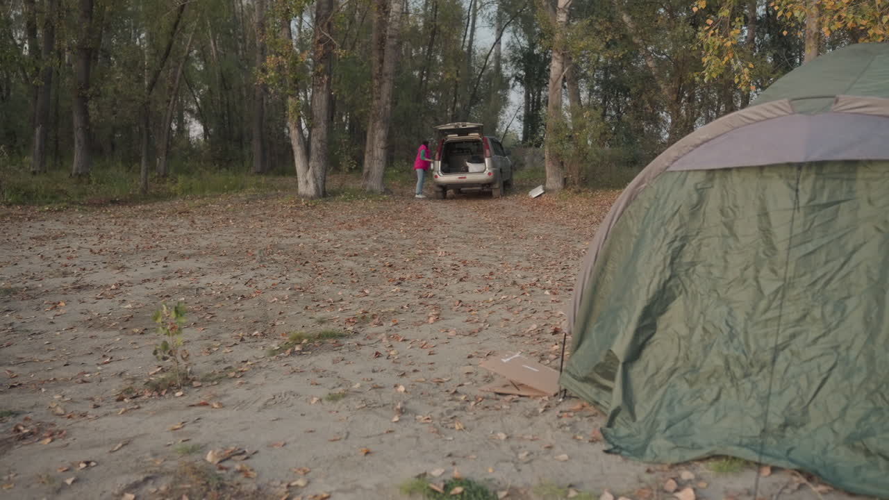 Campingtelt på sandet lysning nær køretøj, camper læsser udstyr ud fra bagagerummet, birketræer i baggrunden, spredte blade på jorden, overskyet lys, stille efterårsstemning og praktisk lejrplads.