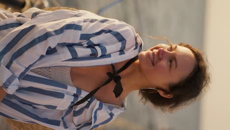 Vertical-video-portrait-of-a-brown-haired-girl-with-large-earrings-who-is-dressed-in-a-white-blue-shirt-and-a-black-bow.-Happy-brown-haired-girl-smiles-and-looks-at-the-camera-against-of-the-sea