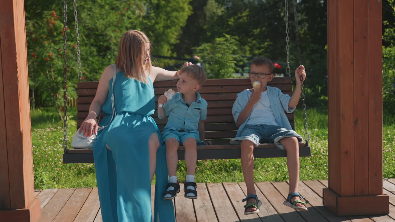 Hvid mor sidder med sønner på træbænk i park. Turkis kjole, to drenge spiser isvafler, solrig sommerdag, blid samtale og latter, afslappet omsorgsøjeblik under familie