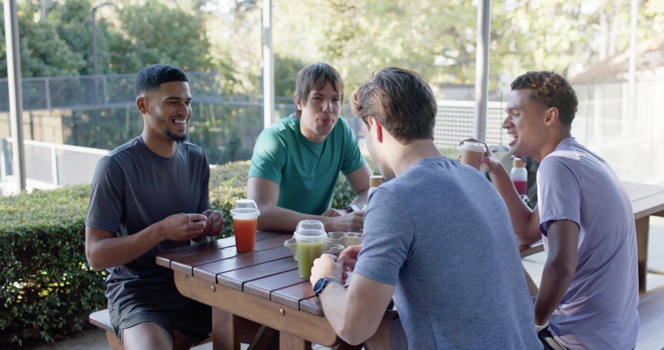 Diverse male friends talking as green-tee starting socializing over to-go cups at patio near court