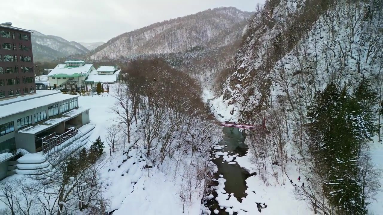 Malownicze ujęcie z drona przemieszczające się wzdłuż rzeki ze skałami pokrytymi śniegiem w kierunku mostu wiszącego Futami w Jozankei Onsen, Hokkaido.