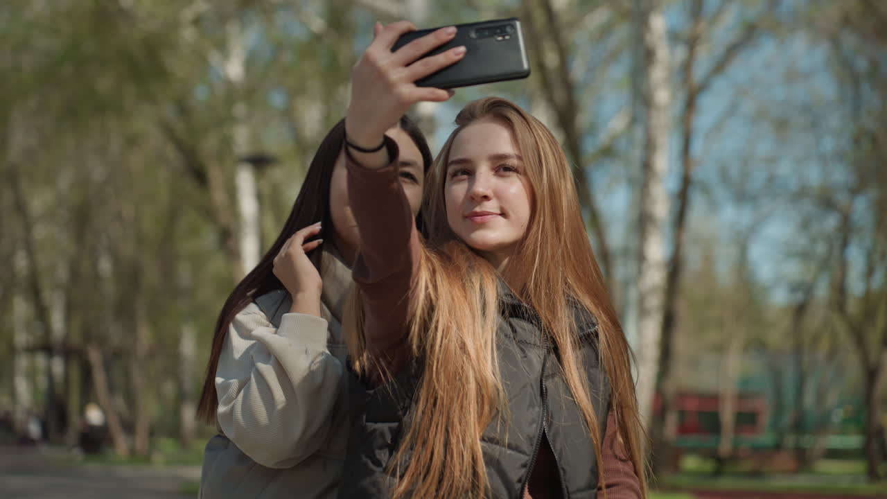 Две женщины позируют для селфи с телефоном, азиатская и белая студентки наклонились друг к другу, смартфон поднят высоко, улыбающиеся лица, парковая дорожка и естественное освещение, атмосфера создателя контента и искренняя энергетика