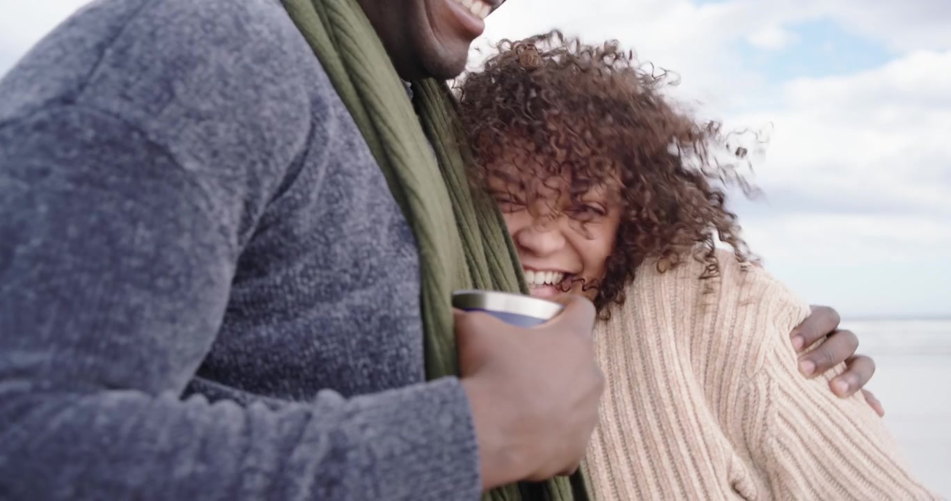 Par af afrikansk afstamning på stranden, mand holder rejsekop, vinden løfter hendes hår, hun putter sig varmt.