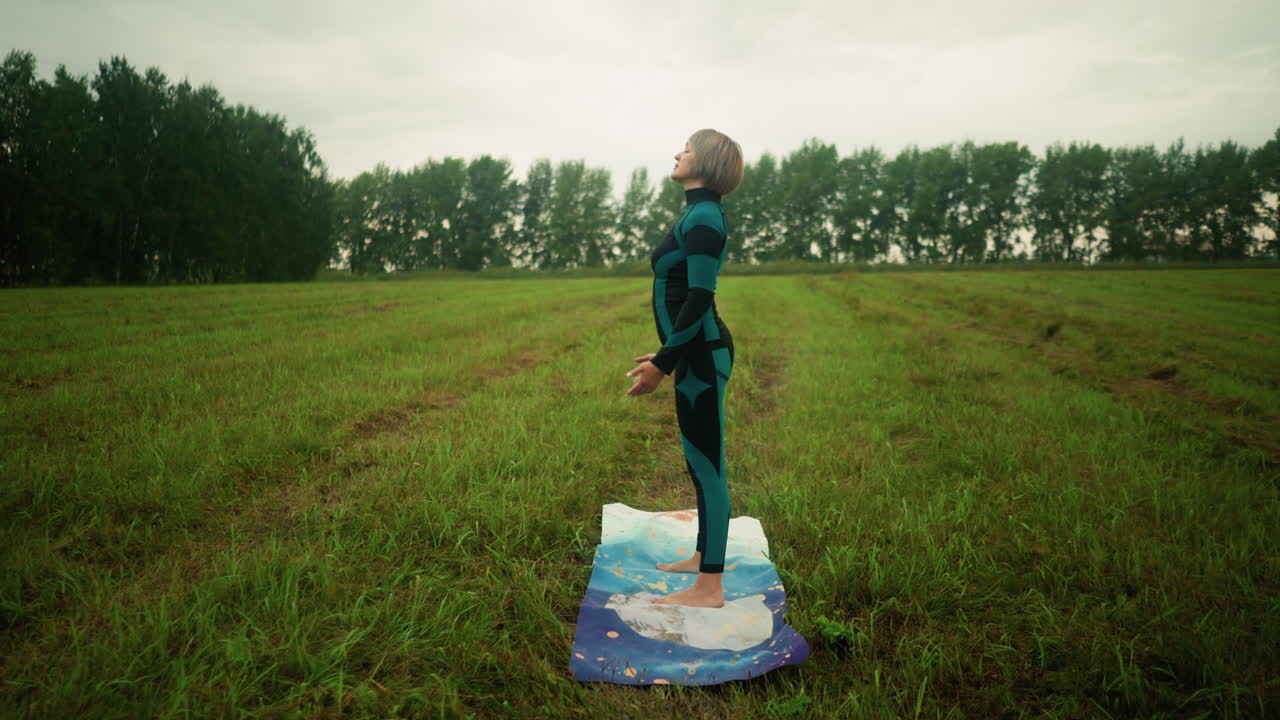 잔디에 있는 매트 위에서 요가를 하고 있는 양복과 레을 입은 여성의 측면 전망, 눈을 감고 천천히 위아래로 움직이는 손, 구름이 가득한 하늘 아래 열린 과 멀리 있는 나무로 둘러싸인 모습