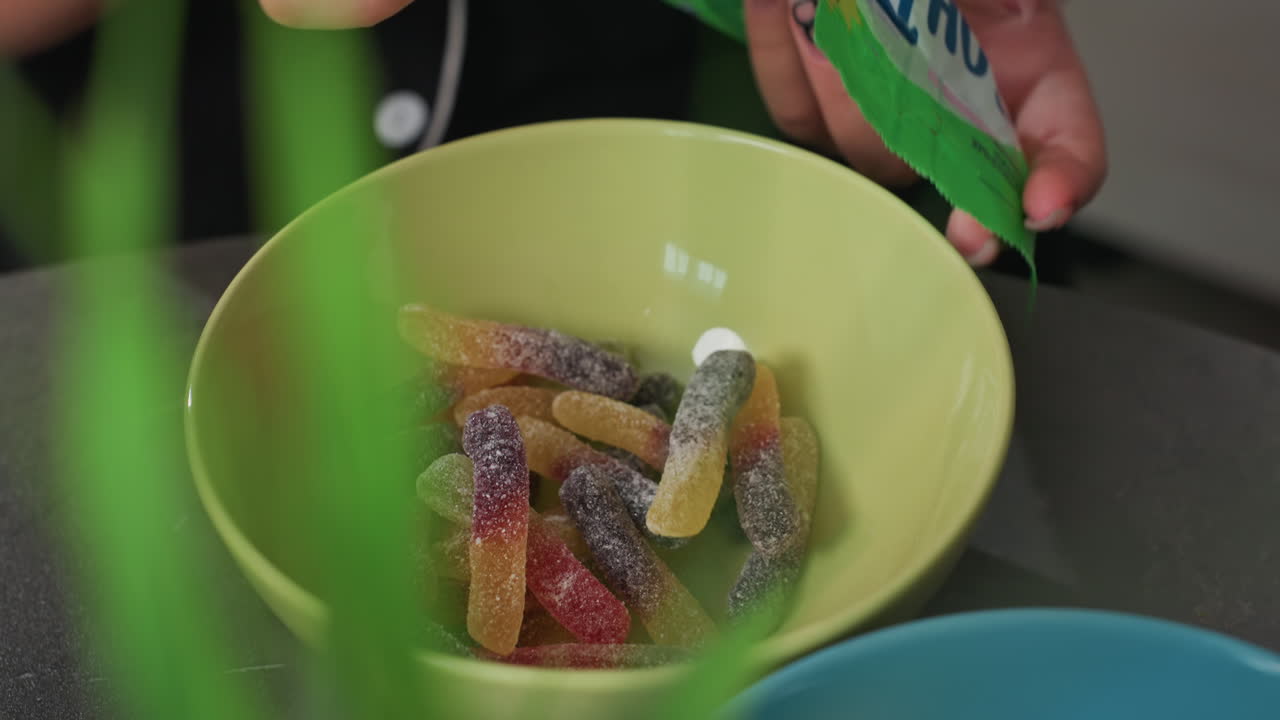 close up colorful gummy candies in yellow bowl with people taking candies, mix of red, yellow, orange, purple sugar coated candies, casual indoor snack scene with hands reaching in
