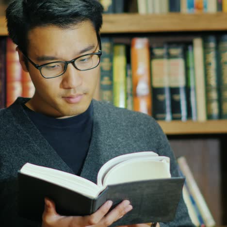 Gut-Aussehender-Asiatischer-Mann-Mit-Brille,-Der-Ein-Buch-In-Der-Bibliothek-Liest