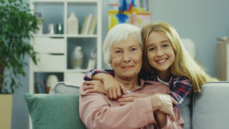 portrait of the happy teenager girl hugging her grandmother while she sitting on the sofa at home and they smiling to the camera