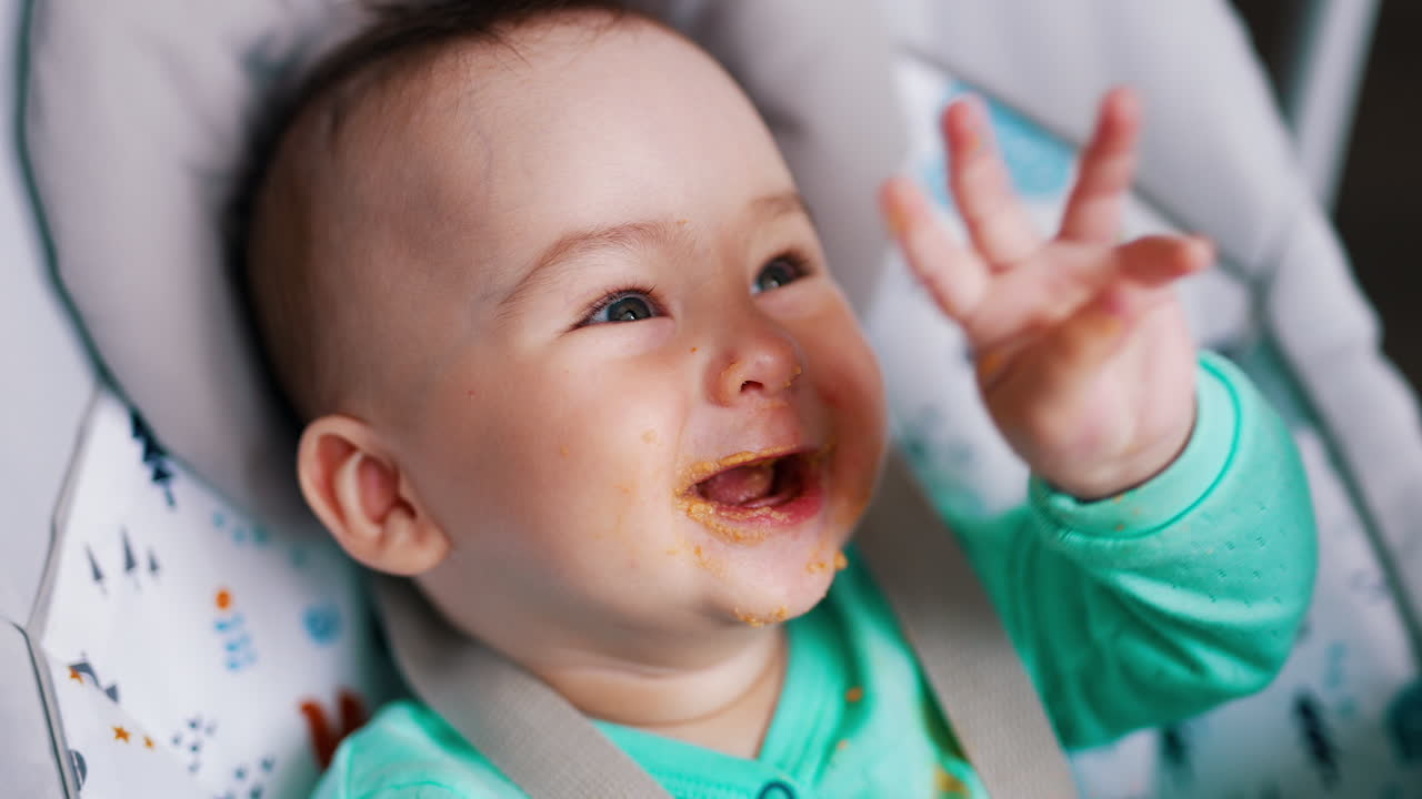 Baby is laughing being fed and rises hands cheerfully. Mom's spoon shoves full spoon into kid's mouth. Close up.