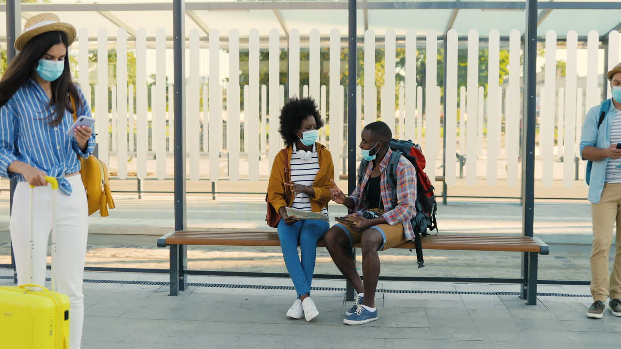 African american young happy man and woman travellers in facial masks sitting at bus stop talking and watching a map and a tablet to plan a route