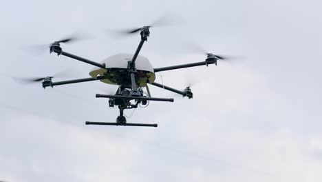 handheld camera shot of drone with six rotors, white body, green circuit board, camera, and two black landing gears in flight against a cloudy sky