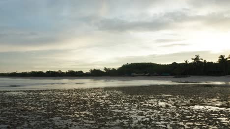 Low-tide-in-tropical-bay-with-silhouette-shore-from-rising-sun-behind-it,-drone