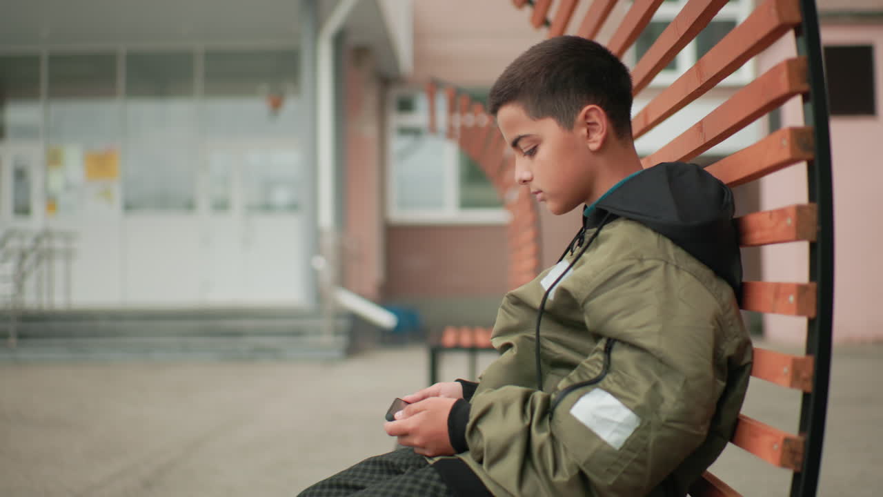 Vista lateral de un niño sentado en un banco con una chaqueta, concentrado en el teléfono jugando a un juego en un patio cerca de la entrada de un edificio escolar bajo una luz suave y diurna, con concentración tranquila y postura relajada.