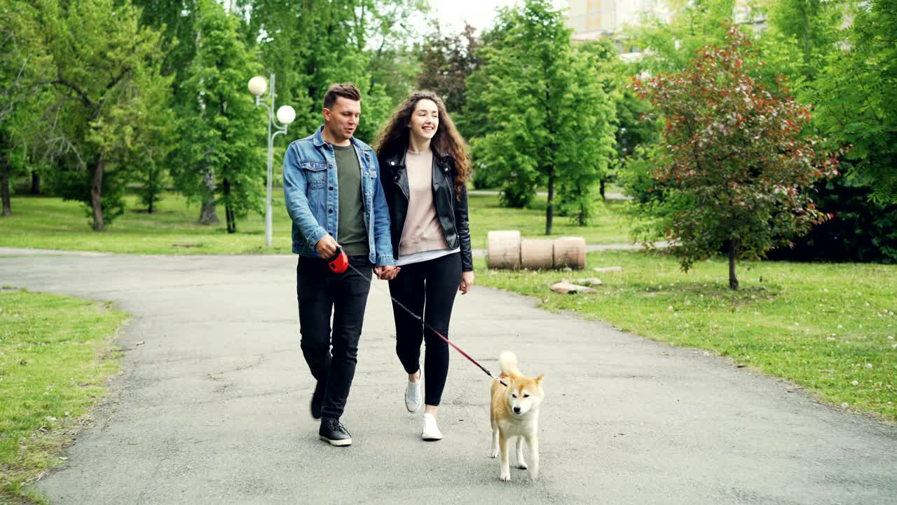 Cheerful guy in denim clothes is walking his dog holding his wife's hand, people and animal are going along the path in park in summer. Active lifestyle concept.