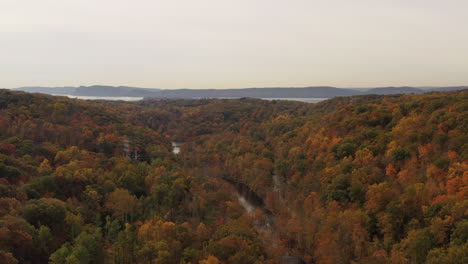 aerial dolly in, on a cloudy day, drone camera tilts down over the stream - tree tops with orange leaves at the new croton dam in westchester county, ny