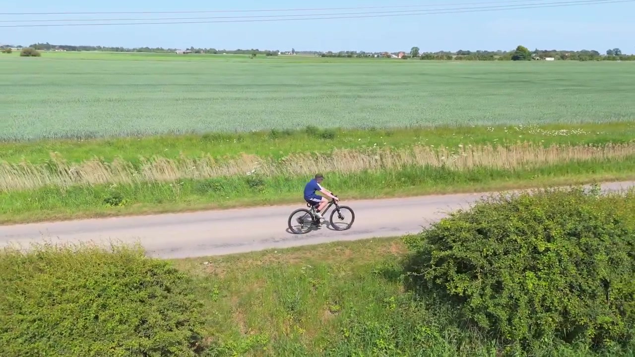 Cyclist rides a bicycle along a country road. The rider wears blue, moving smoothly through green bushes. Lush fields stretch under a bright, clear sky. The cyclist appears relaxed and happy