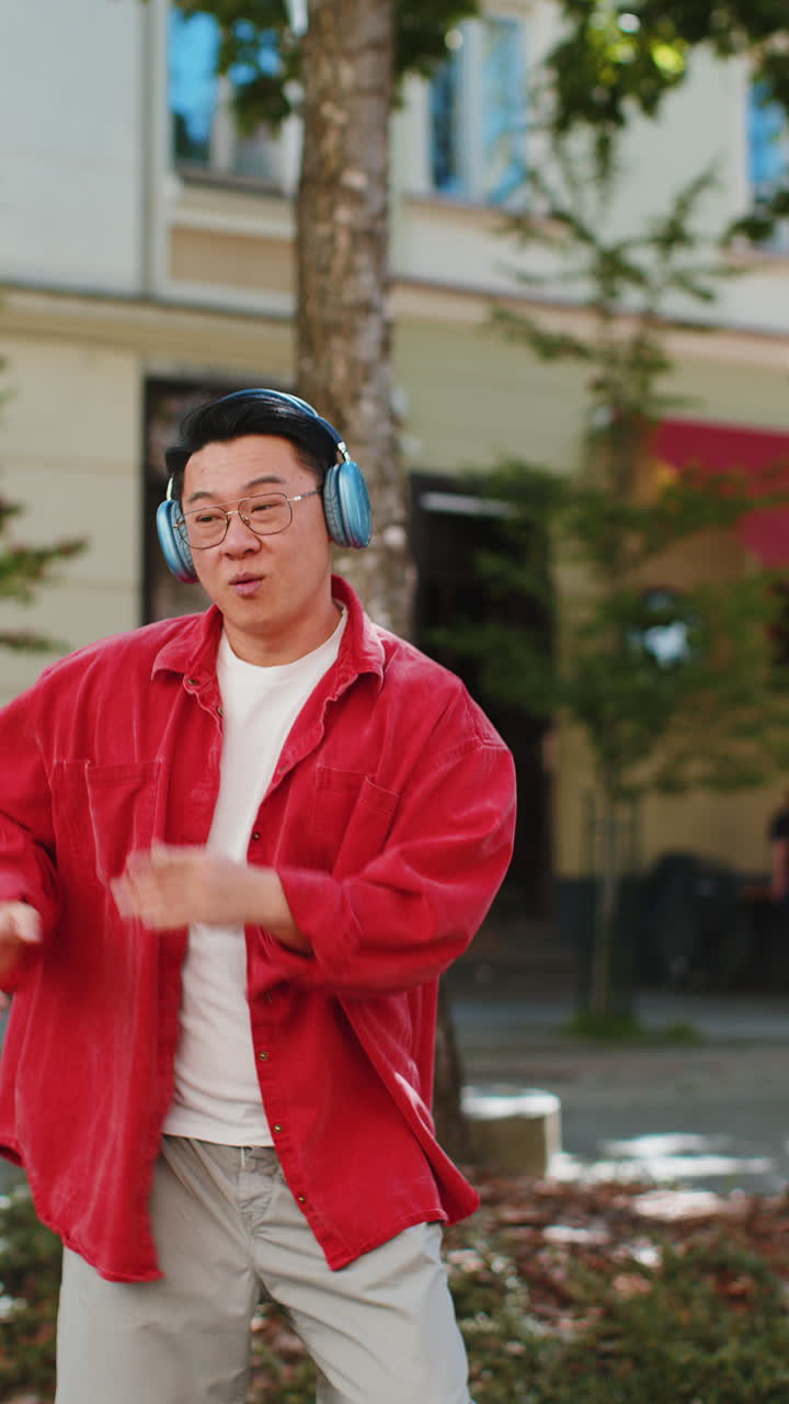 Happy Asian man dancing in wireless headphones listening favorite energetic music on city street