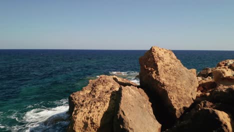 Aerial-dolly-shot-of-a-seashore-with-beautiful-turquoise-water-of-the-Mediterranean-Sea-splashing-on-the-red-coloured-rocks